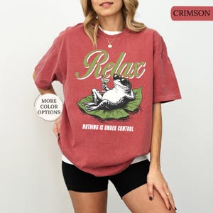 May include: Crimson-colored t-shirt featuring a frog relaxing on a lily pad, holding a cocktail. The word "Relax" is written in green script above the frog, with "Nothing is Under Control" below. The shirt has short sleeves.