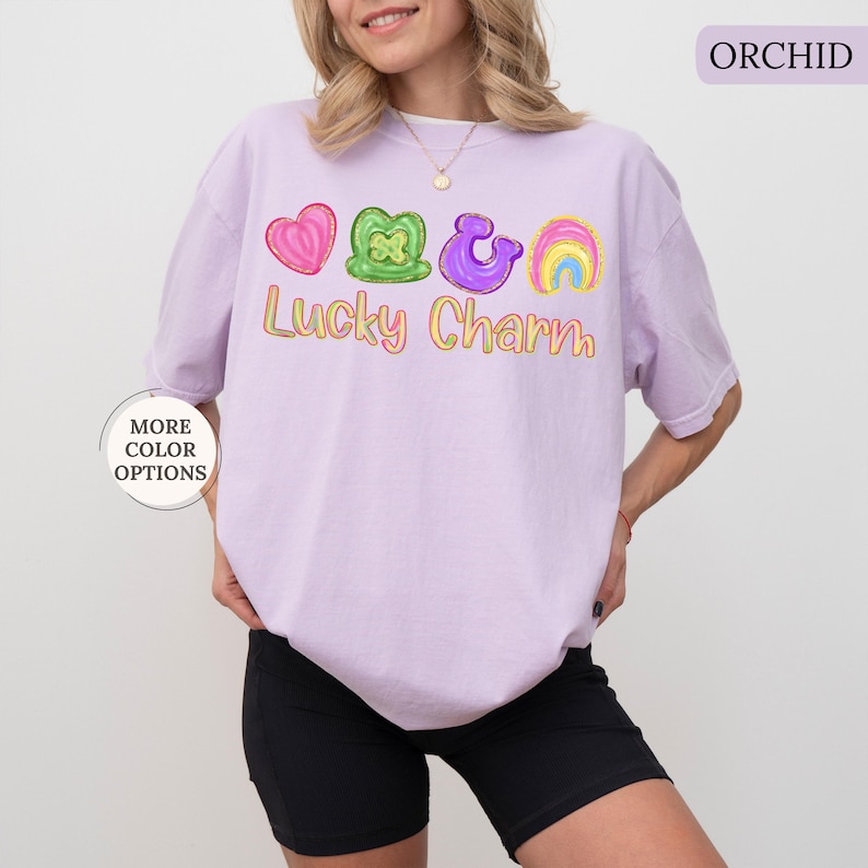 May include: A light purple t-shirt with the text "Lucky Charm" in yellow and a graphic of a heart, shamrock, horseshoe, and rainbow. The shirt also has the text "MORE COLOR OPTIONS" on the left side.