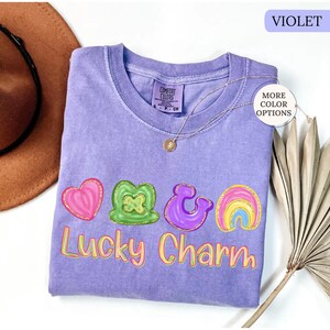 May include: A violet Comfort Colors t-shirt with a "Lucky Charm" graphic. The graphic includes a pink heart, green shamrock, purple ampersand, and rainbow. A gold necklace with a pendant is on the shirt.