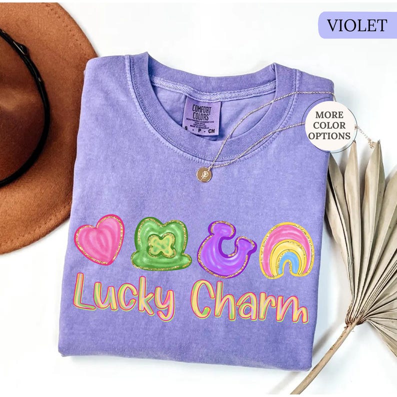 May include: A violet Comfort Colors t-shirt with a "Lucky Charm" graphic. The graphic includes a pink heart, green shamrock, purple ampersand, and rainbow. A gold necklace with a pendant is on the shirt.