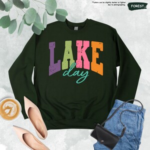 Op de afbeelding: Een bosgroene sweatshirt met de tekst "LAKE day" in een kleurrijke, verweerde lettertype. Het woord "LAKE" is paars, roze en oranje, terwijl "day" turquoise is. De sweatshirt wordt getoond met schoenen, een jeans en een koffiekopje.