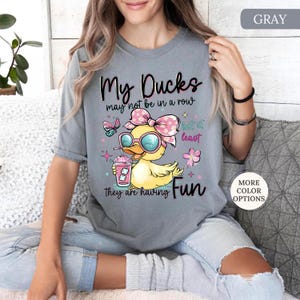 May include: Gray t-shirt with a graphic of a yellow duck wearing sunglasses and a pink bow, holding a pink drink. Text reads "My Ducks may not be in a row but at least they are having Fun."