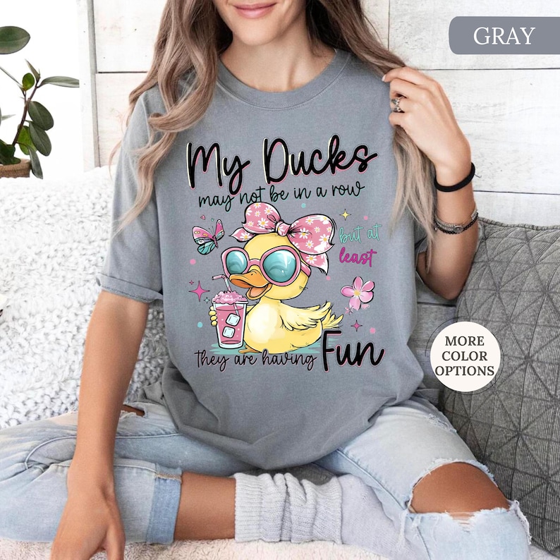 May include: Gray t-shirt with a graphic of a yellow duck wearing sunglasses and a pink bow, holding a pink drink. Text reads "My Ducks may not be in a row but at least they are having Fun."