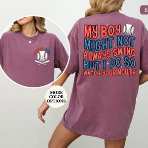 May include: Berry-colored t-shirt with baseball-themed graphics. The front features a baseball and crossed bats with "Baseball Mama" text. The back displays text that reads "My boy might not always swing but I do so watch your mouth" with baseball graphics.