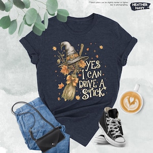 Pode incluir: T-shirt azul marinho mesclado com o texto "YES I CAN DRIVE A STICK" e um gr&aacute;fico de chap&eacute;u de bruxa, vassoura e folhas de outono. A imagem inclui jeans azuis, uma bolsa preta e t&ecirc;nis de cano alto pretos.