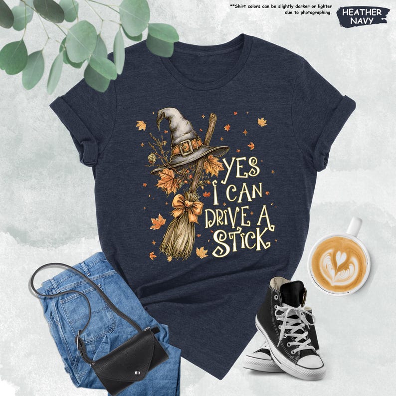 Pode incluir: T-shirt azul marinho mesclado com o texto "YES I CAN DRIVE A STICK" e um gr&aacute;fico de chap&eacute;u de bruxa, vassoura e folhas de outono. A imagem inclui jeans azuis, uma bolsa preta e t&ecirc;nis de cano alto pretos.