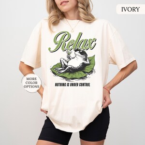 May include: Ivory-colored t-shirt featuring a graphic of a frog relaxing on a lily pad, holding a cocktail. The word "Relax" is arched above the frog, with "Nothing is Under Control" below. The shirt has a relaxed fit.