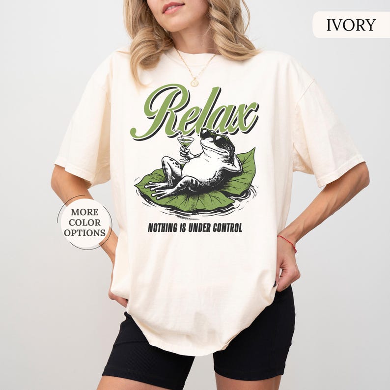 May include: Ivory-colored t-shirt featuring a graphic of a frog relaxing on a lily pad, holding a cocktail. The word "Relax" is arched above the frog, with "Nothing is Under Control" below. The shirt has a relaxed fit.