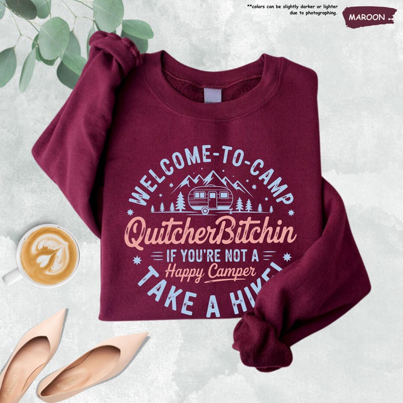 May include: Maroon sweatshirt with the text "WELCOME-TO-CAMP Quitcher Bitchin IF YOU'RE NOT A Happy Camper TAKE A HIVE". The design includes a camper, mountains, and trees. A cup of coffee and a pair of shoes are in the image.