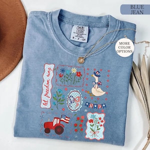 May include: Blue jean colored t-shirt with a patriotic design. The design includes a red tractor, a goose wearing a hat, flowers, and the text "let freedom ring". A gold necklace is draped across the shirt.