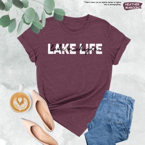 May include: A maroon t-shirt with the text "LAKE MICHIGAN" in white. The shirt is displayed with blue jeans, beige flats, and a cup of coffee. The words "HEATHER MAROON" are in the upper right corner.