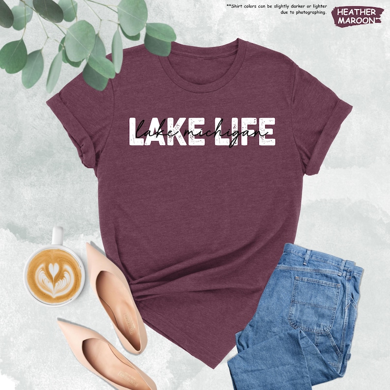 May include: A maroon t-shirt with the text "LAKE MICHIGAN" in white. The shirt is displayed with blue jeans, beige flats, and a cup of coffee. The words "HEATHER MAROON" are in the upper right corner.
