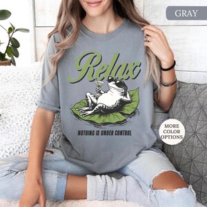 May include: Gray t-shirt featuring a relaxed frog wearing sunglasses and holding a cocktail, lounging on a lily pad. The word "Relax" is above the frog, and "Nothing is Under Control" is below. The shirt has short sleeves.