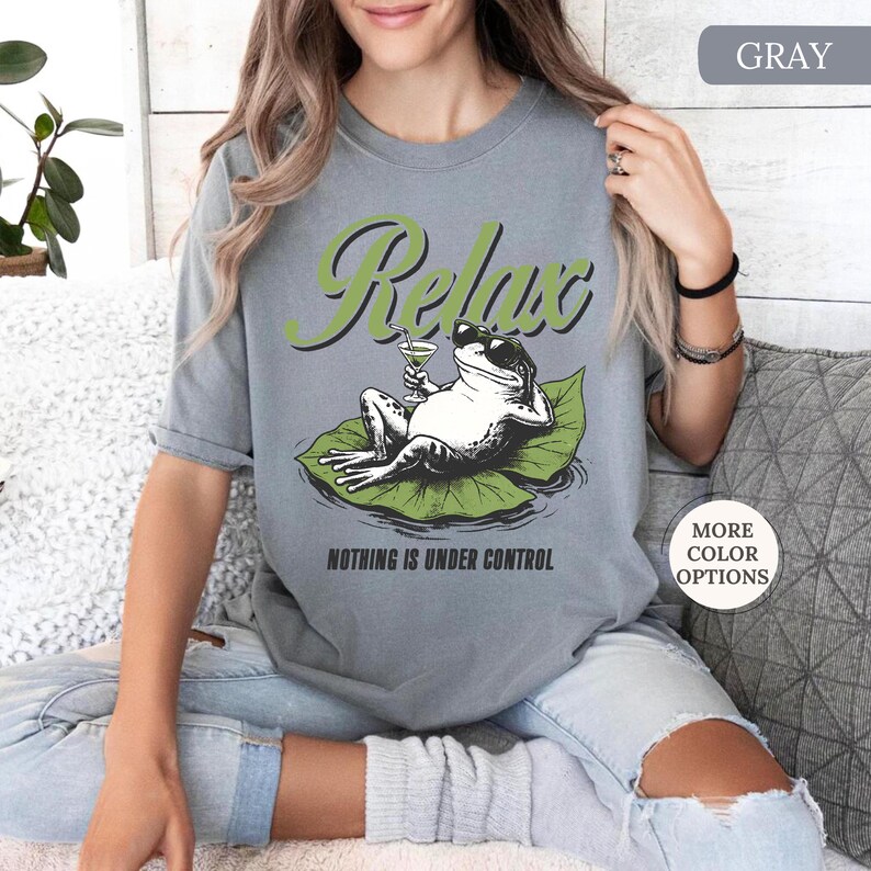 May include: Gray t-shirt featuring a relaxed frog wearing sunglasses and holding a cocktail, lounging on a lily pad. The word "Relax" is above the frog, and "Nothing is Under Control" is below. The shirt has short sleeves.