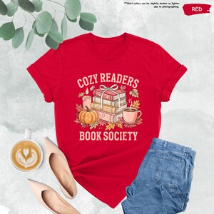 May include: A red t-shirt with the text "COZY READERS BOOK SOCIETY" in white. The graphic includes a stack of books, a pumpkin, a candle, and a cup of coffee. The shirt is paired with blue jeans and beige shoes.