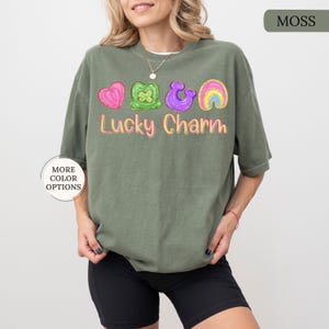 May include: A moss green t-shirt with a colorful "Lucky Charm" graphic. The graphic includes a pink heart, green shamrock, purple horseshoe, and rainbow. The shirt has short sleeves and a relaxed fit.