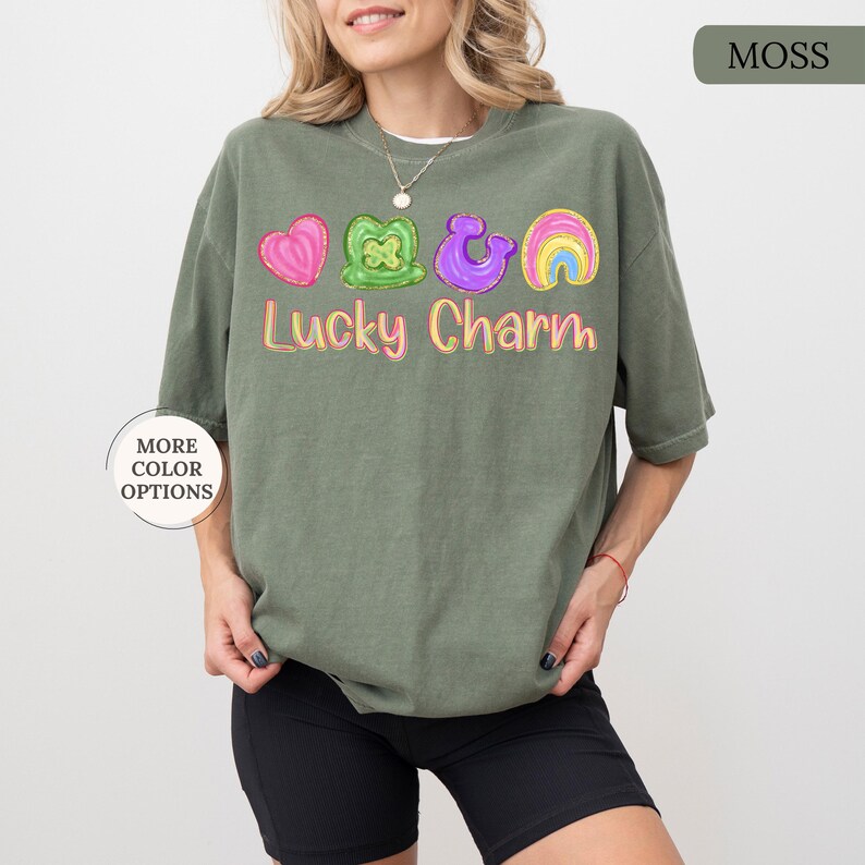 May include: A moss green t-shirt with a colorful "Lucky Charm" graphic. The graphic includes a pink heart, green shamrock, purple horseshoe, and rainbow. The shirt has short sleeves and a relaxed fit.