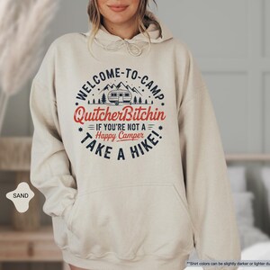 May include: Sand-colored hoodie with a front graphic that reads "WELCOME-TO-CAMP Quitcher Bitchin IF YOU'RE NOT A Happy Camper TAKE A HIKE!" The design includes a camper, trees, and stars. The hoodie has a drawstring hood and a front pocket.