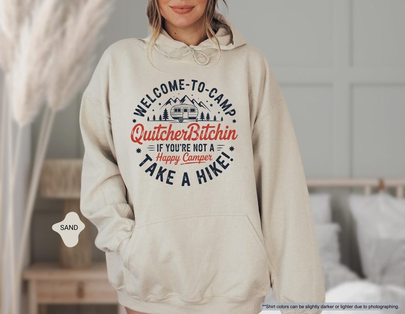May include: Sand-colored hoodie with a front graphic that reads "WELCOME-TO-CAMP Quitcher Bitchin IF YOU'RE NOT A Happy Camper TAKE A HIKE!" The design includes a camper, trees, and stars. The hoodie has a drawstring hood and a front pocket.
