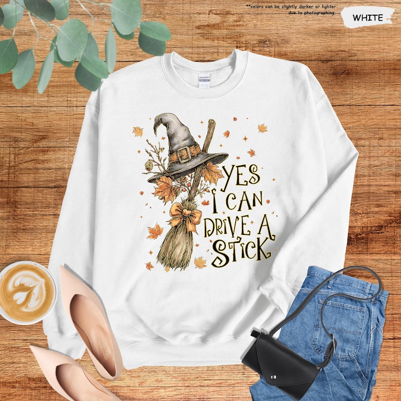 Op de afbeelding: Witte sweatshirt met een heksenhoed, bezem en herfstbladeren. De tekst "YES I CAN DRIVE A STICK" staat op de voorkant. De sweatshirt is van het merk Gildan.