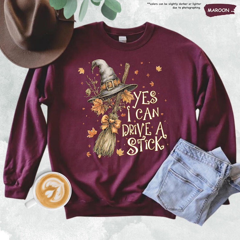 Op de afbeelding: Bordeaux sweatshirt met een heksenhoed, bezem en herfstbladeren. De tekst luidt "YES I CAN DRIVE A STICK". Een bruine hoed, jeans en een kopje koffie zijn ook zichtbaar.