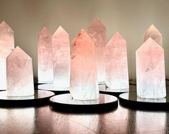 Puede incluir: Una colección de torres de cristal de cuarzo rosa iluminadas. Cada cristal está montado sobre una base circular negra. Los cristales están iluminados desde el interior, creando un efecto suave y brillante con tonos rosas y blancos.