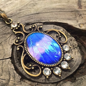 Op de afbeelding: Een vintage-stijl hangerketting met een ovale, iriserende blauw-paarse middensteen. De hanger heeft een sierlijke, bronskleurige metalen zetting, accenten van heldere kristallen en een bijpassende ketting.