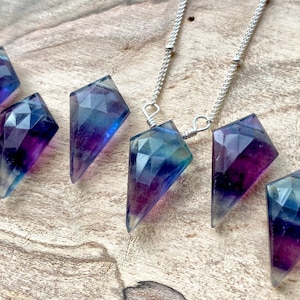 May include: A silver chain necklace with a faceted purple and blue fluorite pendant. The pendant is shaped like a diamond and has a pointed tip. There are several other fluorite pendants of the same shape and color lying on a wooden surface.