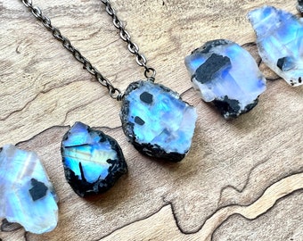 May include: A silver chain necklace with a large, iridescent moonstone pendant. The moonstone is raw and has a black matrix. The necklace is displayed on a wooden surface with other moonstone pieces.