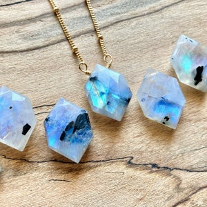 Può includere: Una collana con catena dorata con sei ciondoli in pietra di luna arcobaleno iridescente. Ogni ciondolo è una forma geometrica sfaccettata con inclusiones nere.