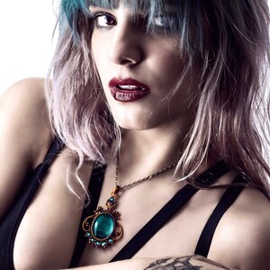 May include: A woman with blue and pink hair wearing a black top with a gold chain necklace featuring a large turquoise gemstone pendant. The woman has a black tattoo on her hand.