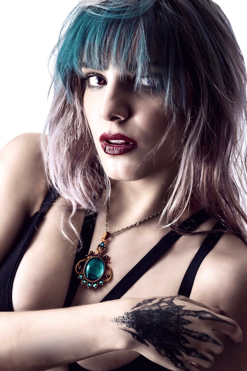 May include: A woman with blue and pink hair wearing a black top with a gold chain necklace featuring a large turquoise gemstone pendant. The woman has a black tattoo on her hand.