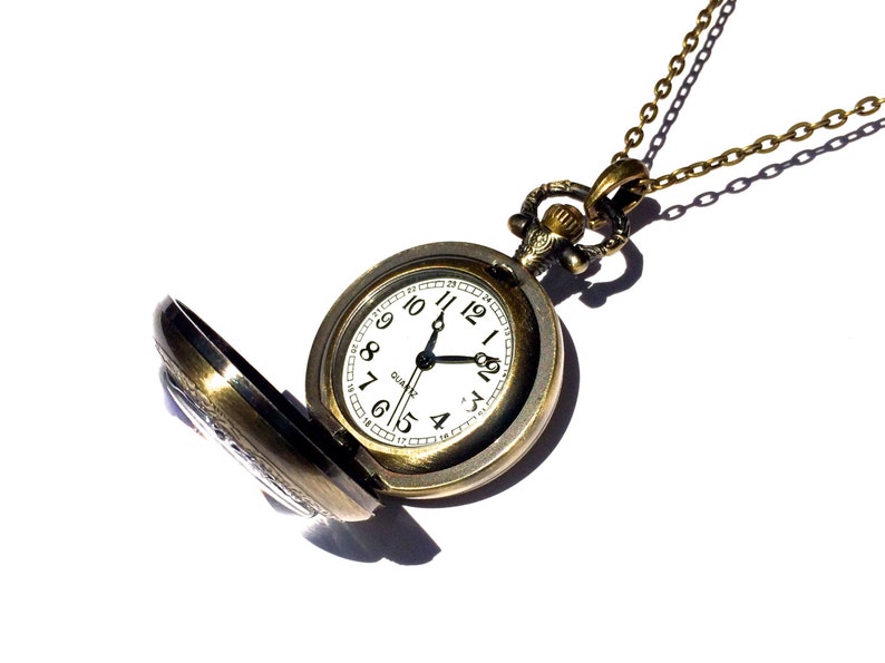 Op de afbeelding: Een gouden zakhorloge met een witte wijzerplaat en zwarte wijzers. Het horloge is open en heeft een ketting eraan.