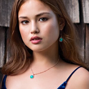 May include: A woman wearing a blue velvet top with a gold chain necklace and earrings featuring a green gemstone pendant.