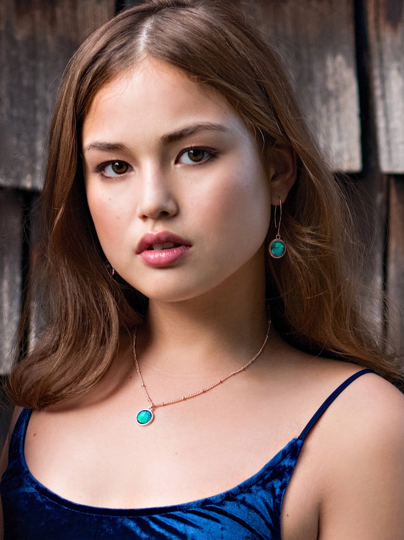 May include: A woman wearing a blue velvet top with a gold chain necklace and earrings featuring a green gemstone pendant.