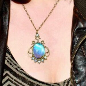 Puede incluir: Un collar de cadena plateada con un gran colgante de piedra preciosa azul iridiscente de forma ovalada engastado en un marco de filigrana dorado.
