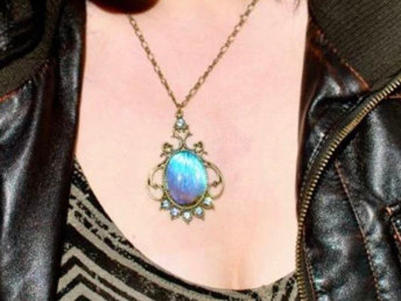 Puede incluir: Un collar de cadena plateada con un gran colgante de piedra preciosa azul iridiscente de forma ovalada engastado en un marco de filigrana dorado.