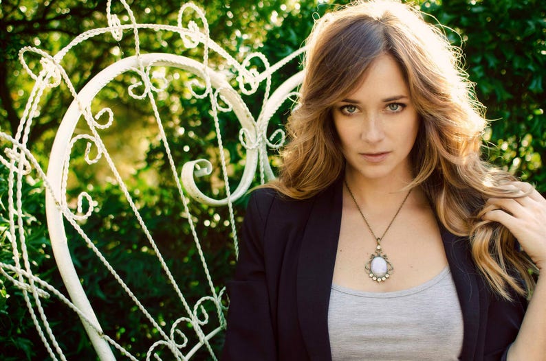 A woman with long, wavy blonde hair stands in front of a white wrought iron gate, wearing a black blazer and a necklace with a pendant.