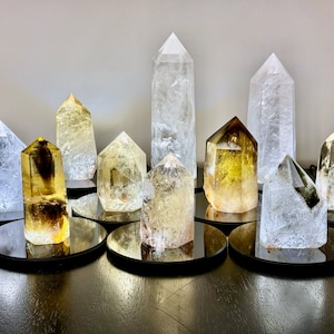 May include: A collection of clear and golden quartz crystal points on mirrored bases. The crystals are arranged in a circular pattern on a dark wooden surface.