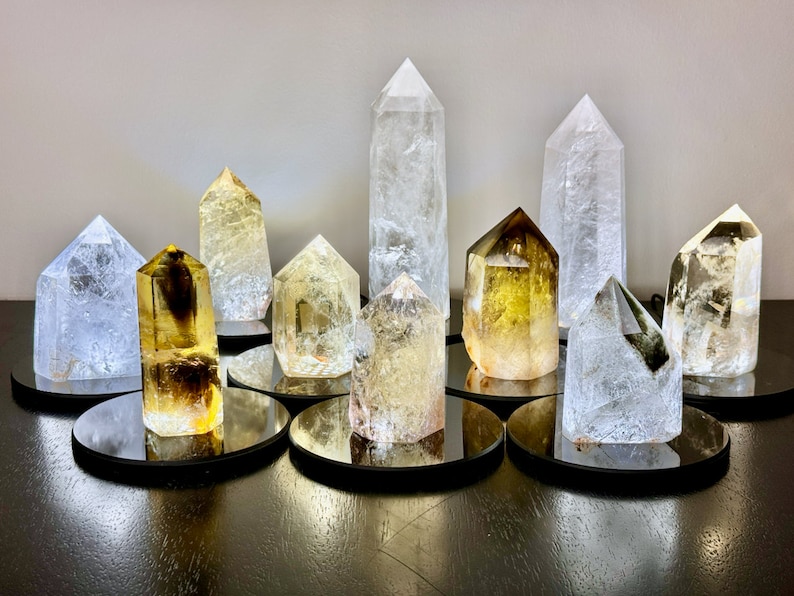 May include: A collection of clear and golden quartz crystal points on mirrored bases. The crystals are arranged in a circular pattern on a dark wooden surface.