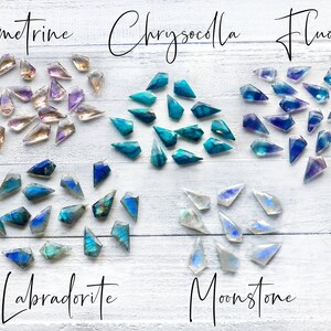 May include: Assortment of faceted gemstone points in various colors, including amethyst, chrysocolla, fluorite, labradorite, and moonstone. The gemstones are arranged in rows on a white wooden surface.