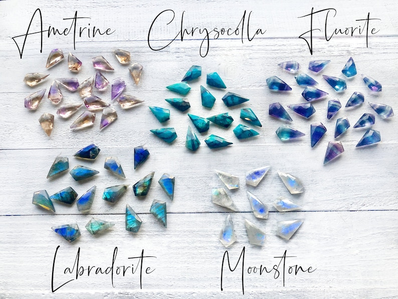 May include: Assortment of faceted gemstone points in various colors, including amethyst, chrysocolla, fluorite, labradorite, and moonstone. The gemstones are arranged in rows on a white wooden surface.