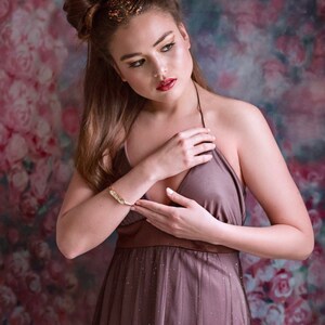 May include: A woman with brown hair styled in two buns wears a mauve sparkly dress with a halter neckline. She is posing with her hands clasped in front of her chest. The background is a blurred floral pattern.