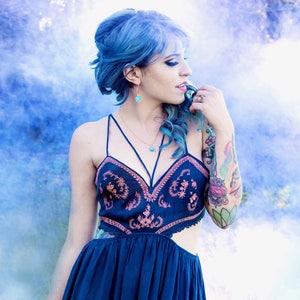 May include: A woman with blue hair wearing a dark blue dress with embroidery and a gold necklace. She has a tattoo on her arm and is standing in front of a blue and white smoke background.