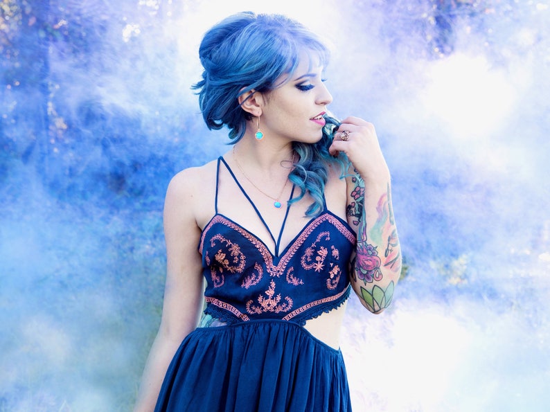May include: A woman with blue hair wearing a dark blue dress with embroidery and a gold necklace. She has a tattoo on her arm and is standing in front of a blue and white smoke background.