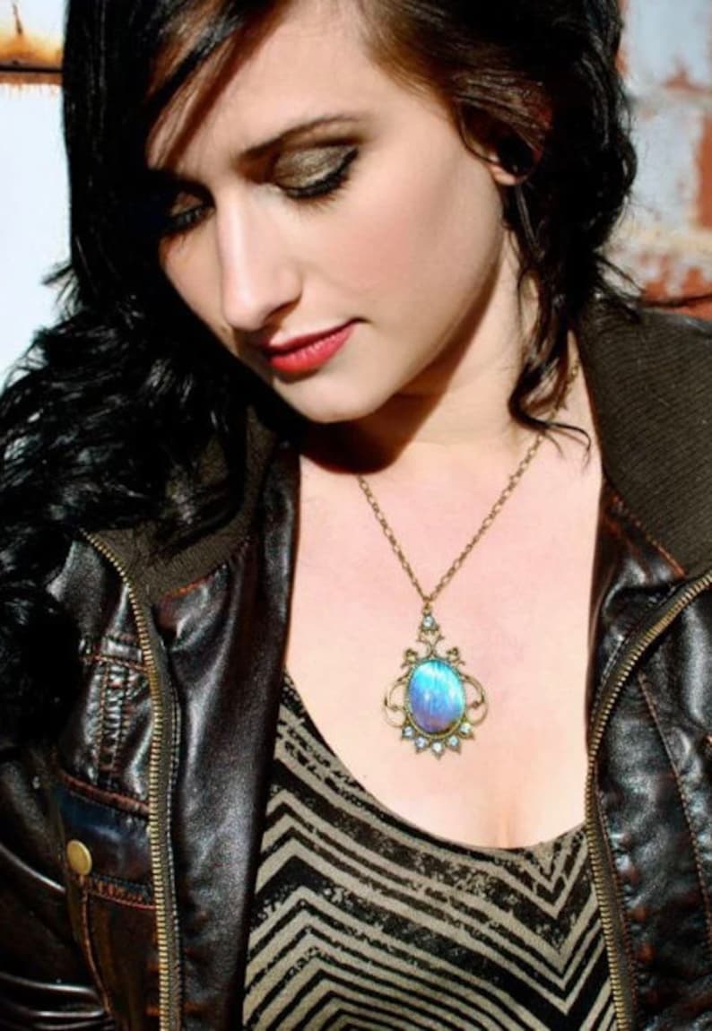 Puede incluir: Una mujer lleva una chaqueta de cuero marr&oacute;n con cremallera y un collar con un colgante de piedra preciosa azul y morado. El collar tiene una cadena dorada y un dise&ntilde;o met&aacute;lico detallado.