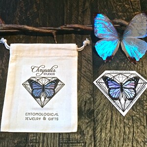 K&ouml;nnte beinhalten: Eine wei&szlig;e Baumwolltasche mit Kordelzug, verziert mit einem Schmetterlingsmotiv und dem Text "Chrysalis Studios" und "Entomological Jewelry & Gifts". Ein blauer Schmetterling liegt auf einer Holzoberfl&auml;che neben der Tasche.