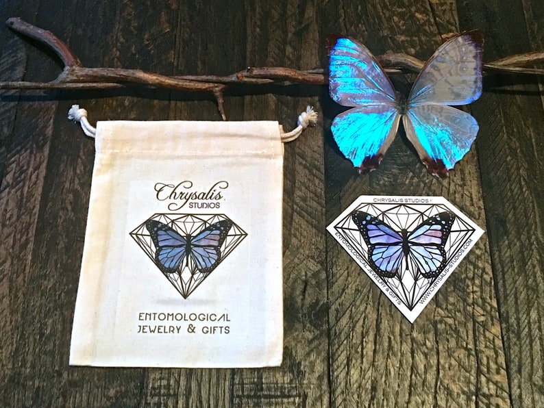 K&ouml;nnte beinhalten: Eine wei&szlig;e Baumwolltasche mit Kordelzug, verziert mit einem Schmetterlingsmotiv und dem Text "Chrysalis Studios" und "Entomological Jewelry & Gifts". Ein blauer Schmetterling liegt auf einer Holzoberfl&auml;che neben der Tasche.