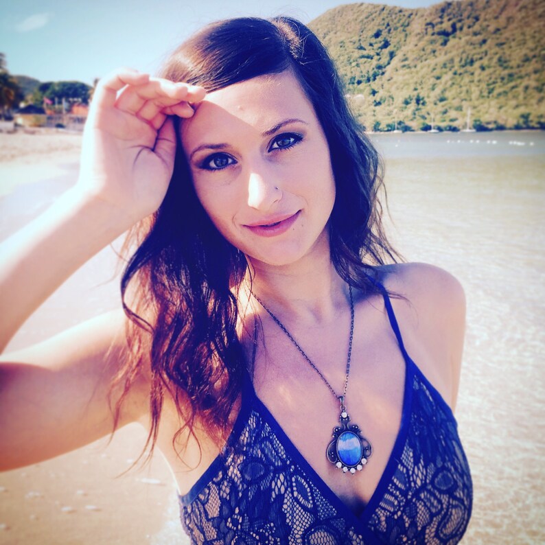 Op de afbeelding: Een vrouw draagt een donkerblauwe kanten top en een hangerketting met een grote blauwe steen. De ketting heeft een sierlijke, antieke setting. De achtergrond toont een strand en de oceaan met een berg in de verte.