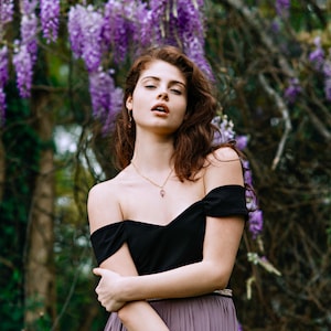 May include: A woman with long brown hair wearing a black off-the-shoulder top and a purple skirt stands in front of a wall of purple wisteria flowers.
